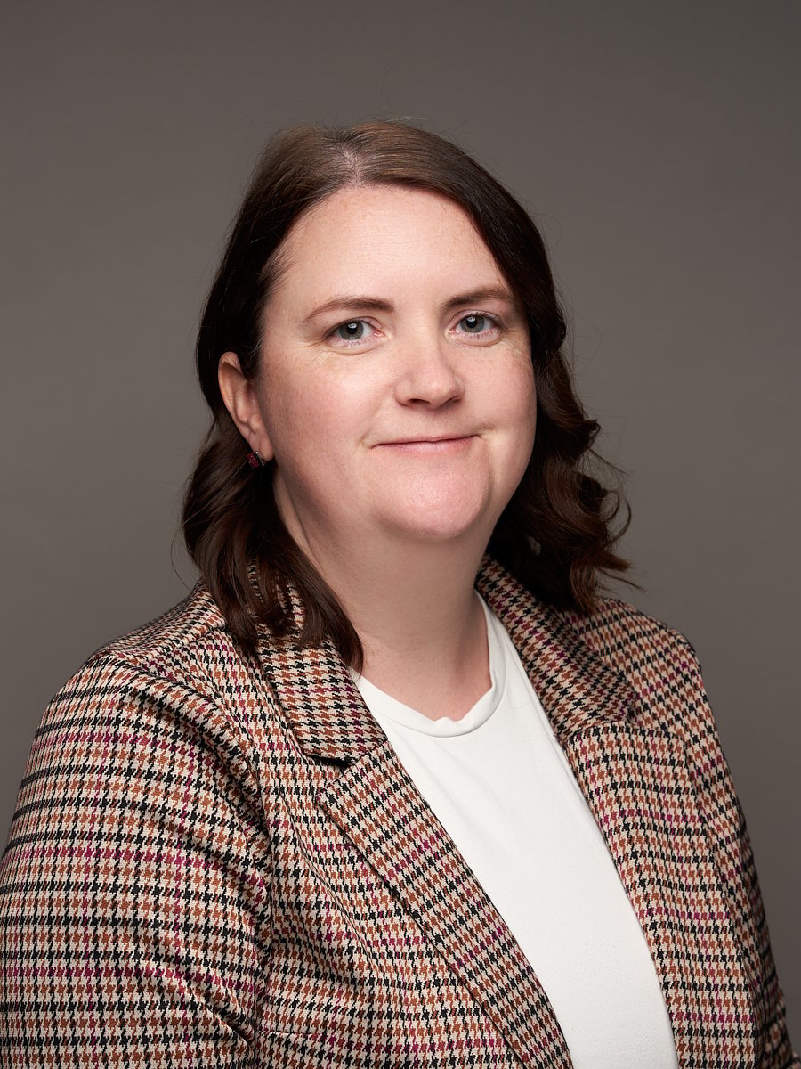 A woman with wavy brown hair and plaid suit jacket against a grey background.
