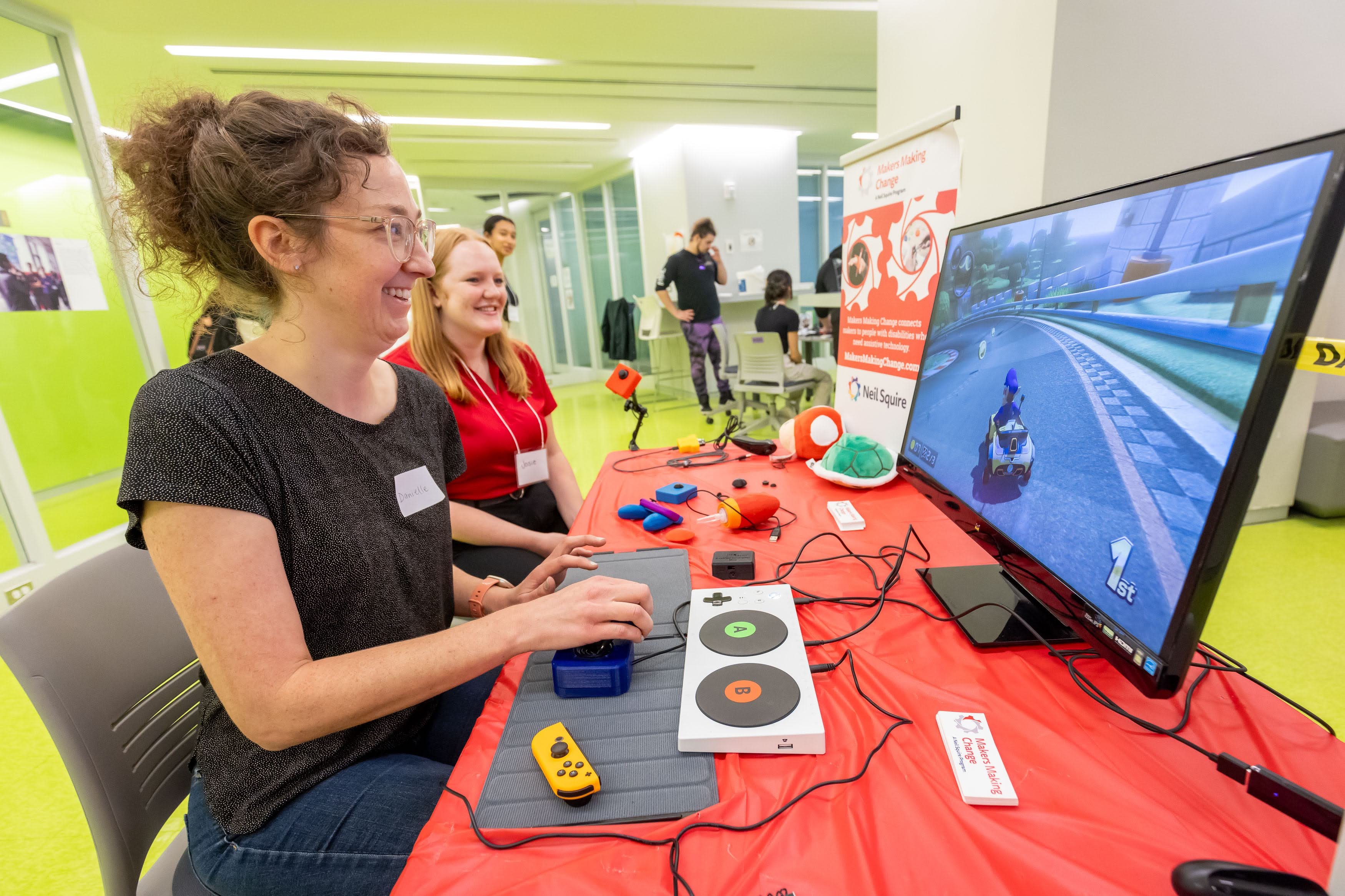 A smiling event participant using a joystick and assistive switch connected to an Xbox Adaptive Controller to play Mario kart. The adaptive gaming gear is sitting on a table with a red tablecloth and other event participants can be seen in the background.