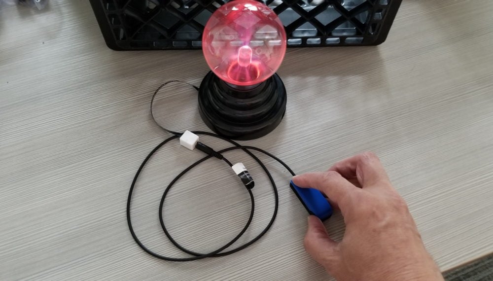 A hand pressing on a 3d printed assistive switch that's plugged into a plasma globe toy via a battery interrupter. The globe is glowing bright pink.
