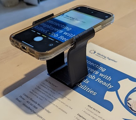 A black 3d printed magnification stand holding an iphone. The camera app is opened and zoomed in on a paper underneath.