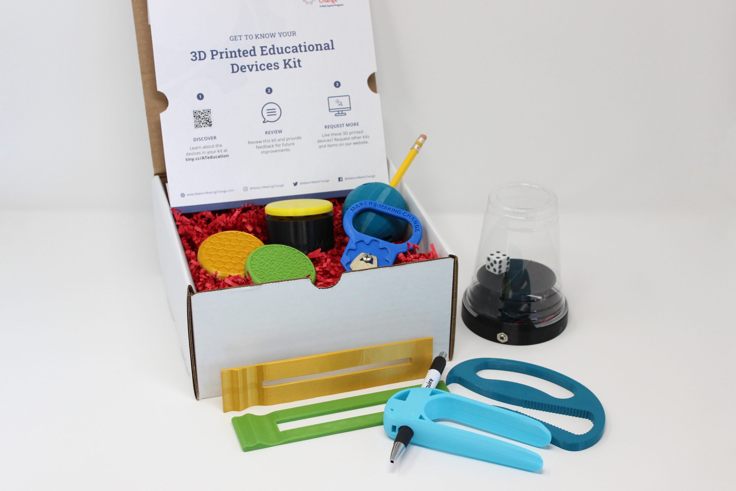 A box of gold, green, and blue 3D printed writing aids, dyslexia reading bars, key turner, bottle opener, acecss switch, and a dice spinner