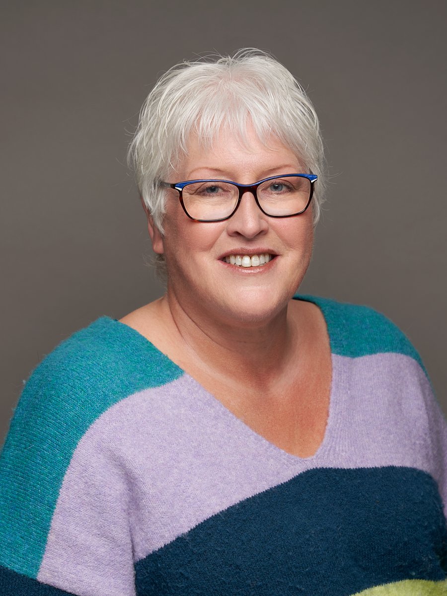 A women with white hair, blue rimmed glasses, and a blue and purple sweater against a grey background.