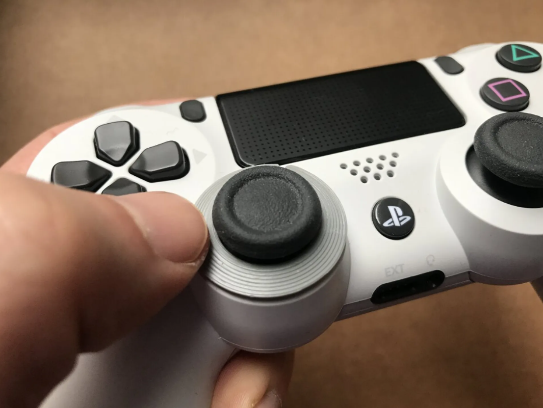 A thumb resting on a grey 3d printed ring attached to the left thumbstick of a PS4 controller.