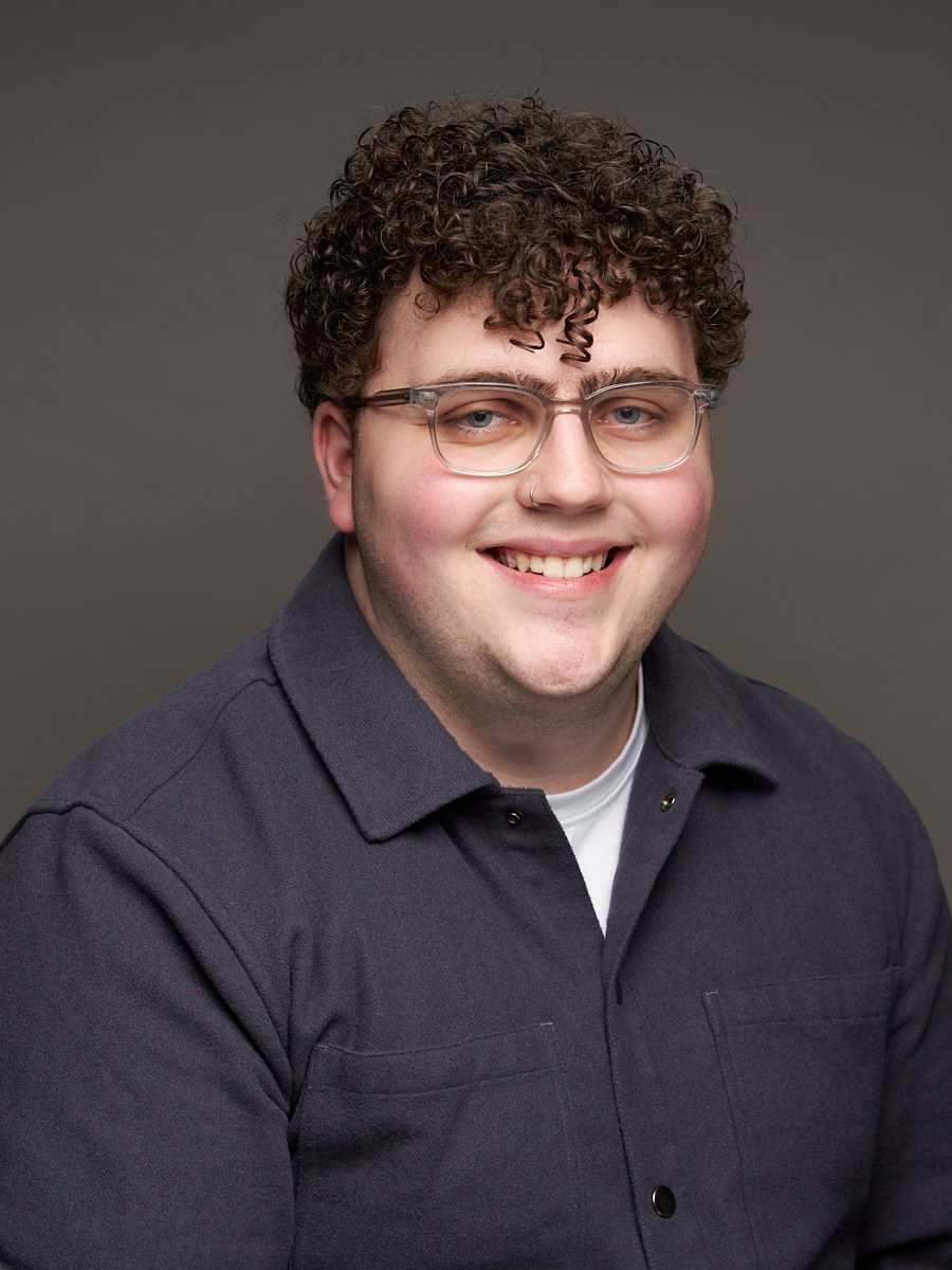 A man with curly brown hair, glasses and dark grey button up against a grey background.