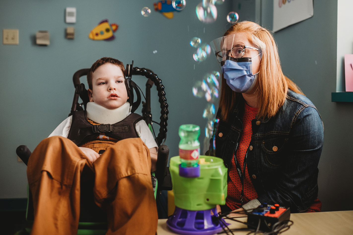 A young boy in a wheelchair looking at bubbles in the air. To the right is his occupational therapist operating the bubble blower