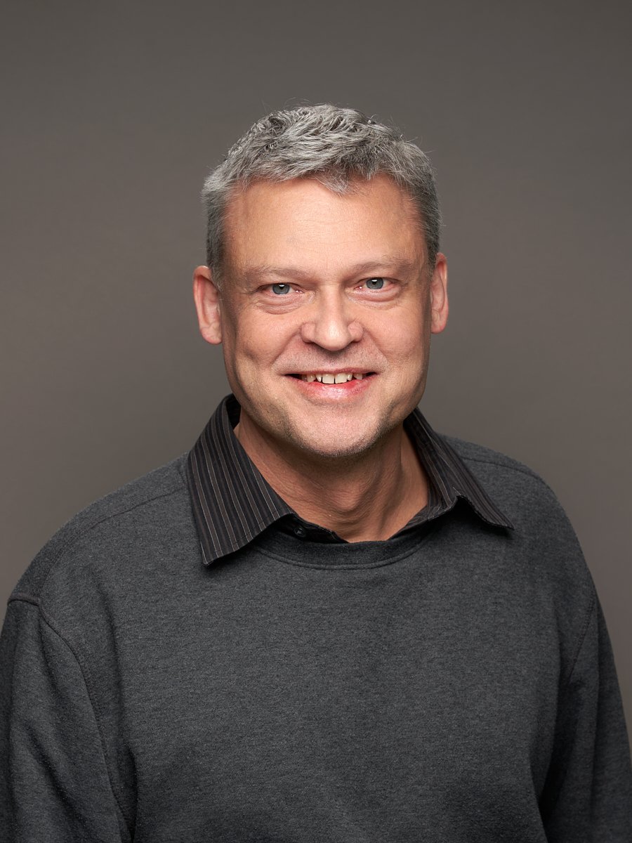 A man with grey hair and grey sweater against a dark background.