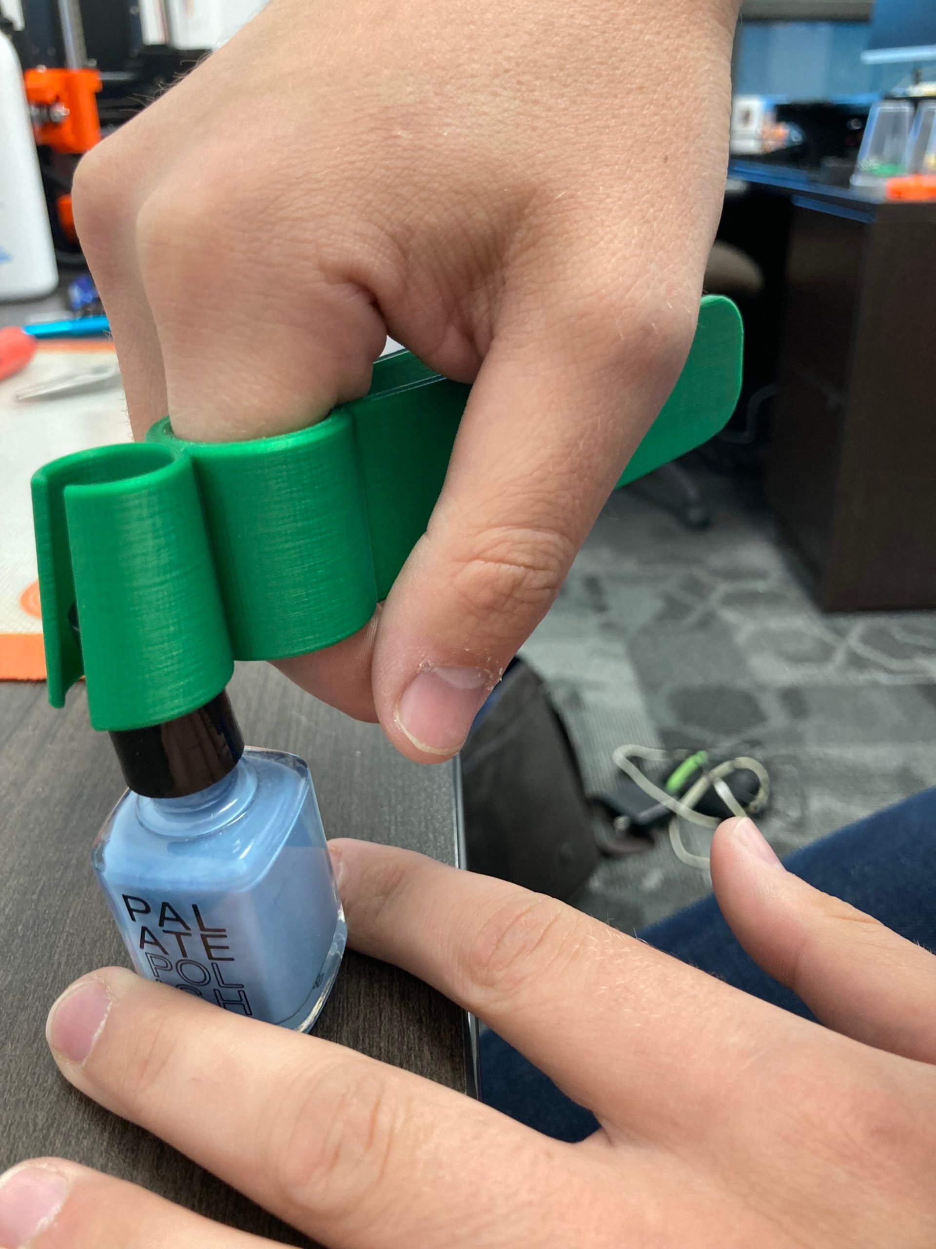 A hand using the handle of a green 3d printed assistive nail polish applicator to grip the lid to a blue nail polish