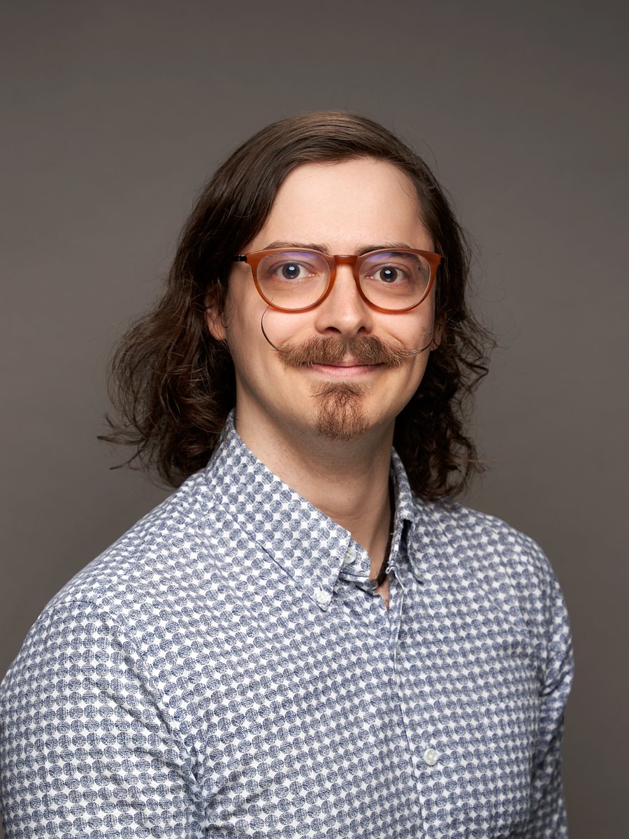 A man with long brown hair and moustache wearing a blue checkered button up against a grey background.
