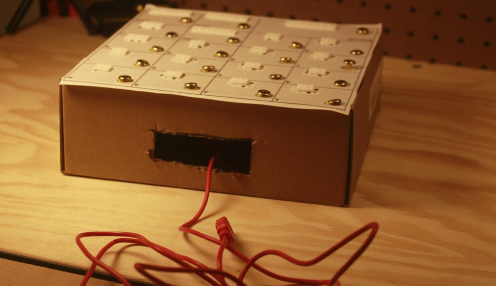 An assembledbraille calculator with metal pins and 3d printed labels on a cardboard box. There is a rectangular hole cut out with a red alligator clip coming out.