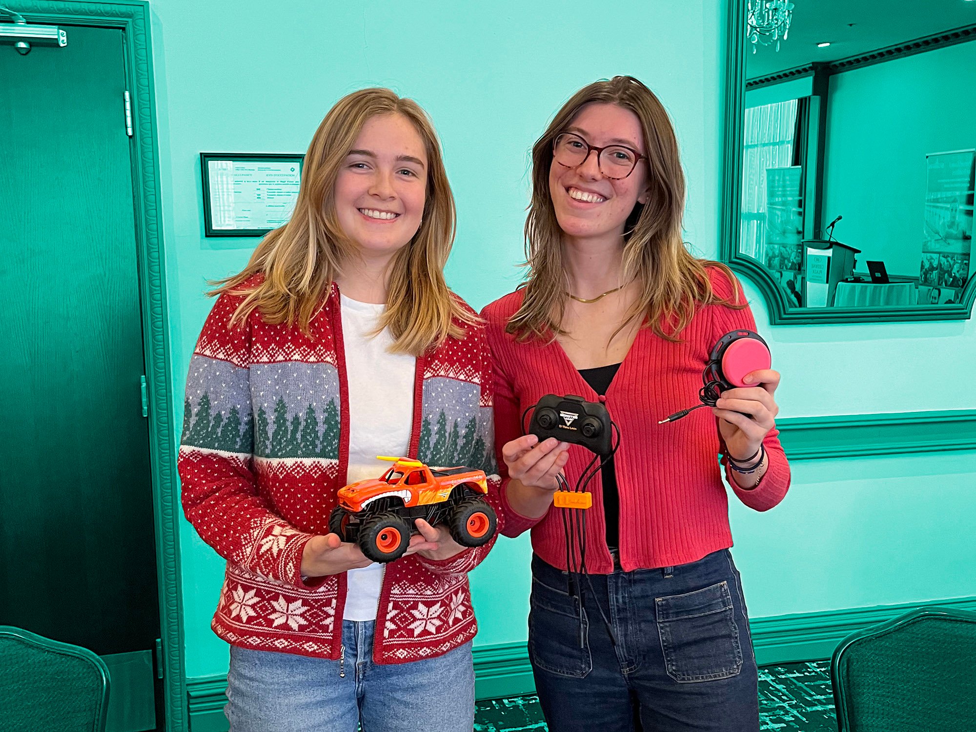 Two woman dressed in festive sweaters holding an adapted car and switch