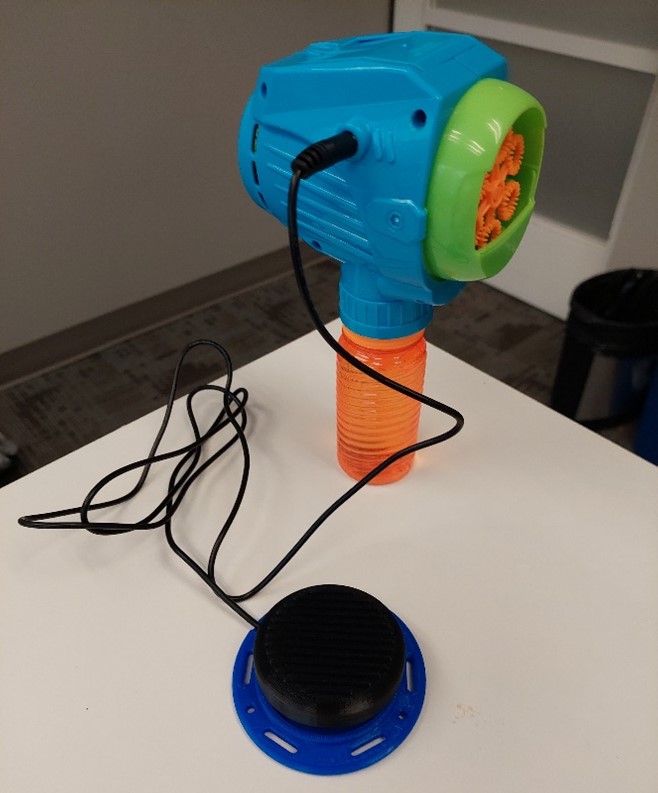 A blue, green, and orange switch adapted bubble blower with a black 3d printed assistive switch plugged in.