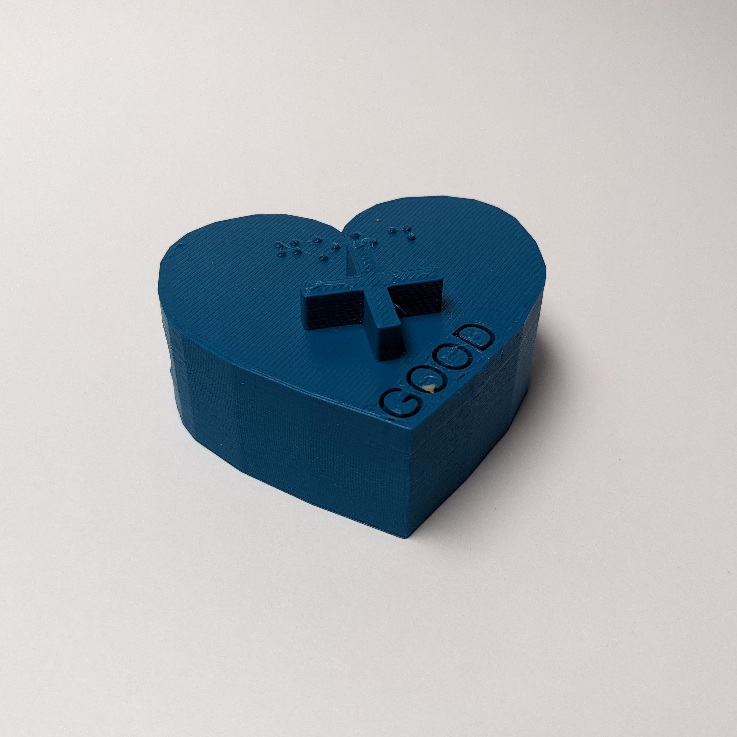 A blue 3d printed project core symbol of the 'Good' symbol against a white surface. The symbol consists of a blue heart shaped base with a smooth textured side. On the top of the symbol, the word 'Good' is engraved, 'Good' is written in Braille, and there is a raised cross or plus symbol.