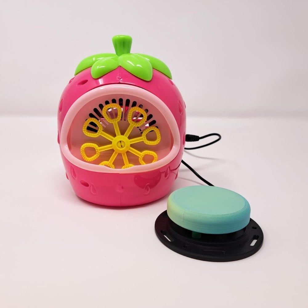 A pink strawberry bubble machine that has been switch adapted to work with an assistive switch. A large green assistive switch is plugged into it.