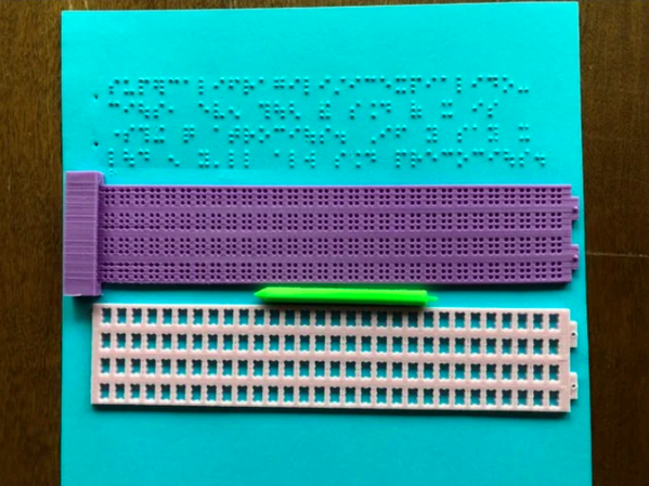 A pink and purple Braille slate with a green stylus in front of a blue sheet of paper with Braille writing