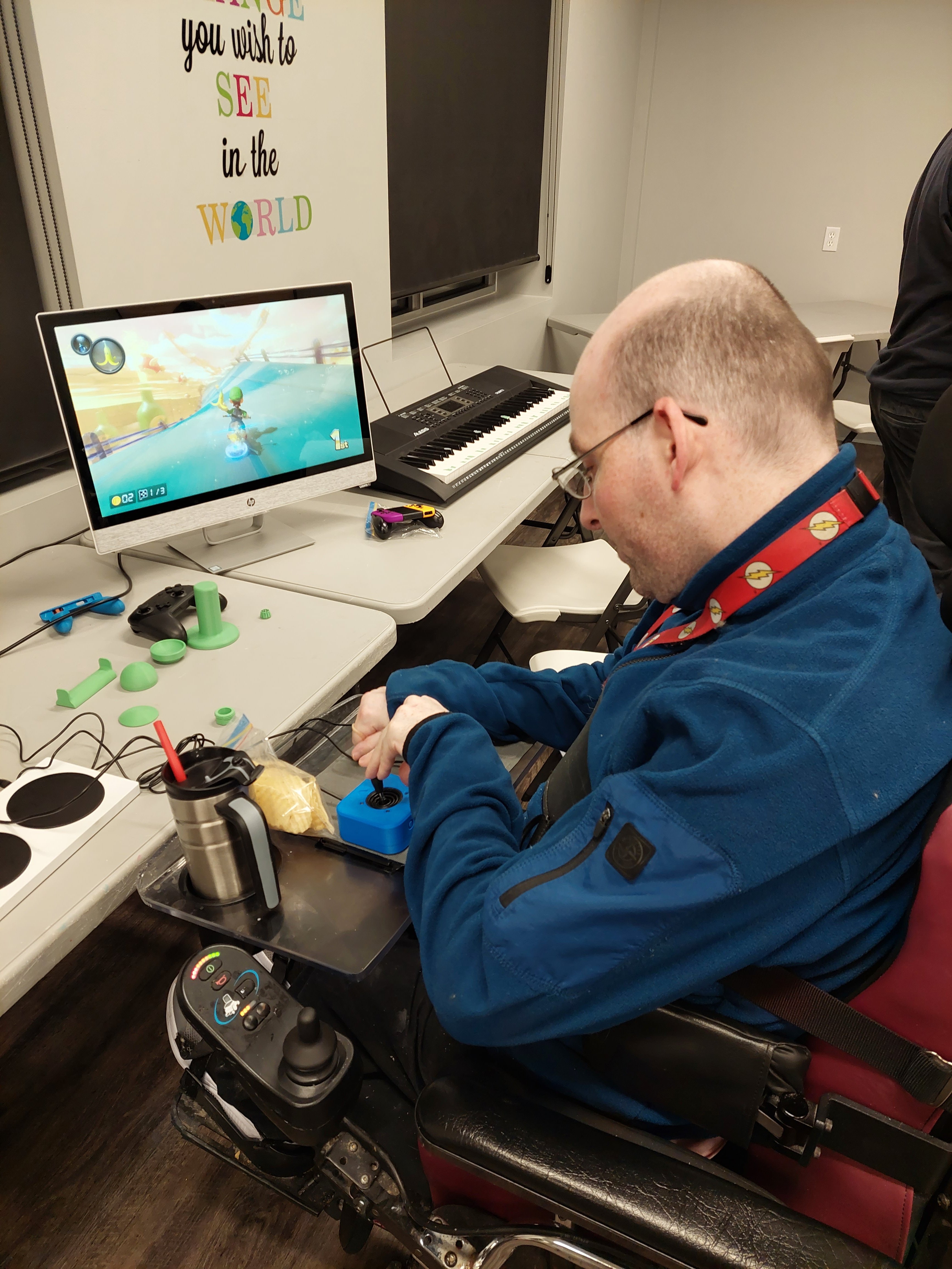 person in a wheelchair using an adaptive joystick with both hands to steer in Mario Kart displayed on a computer monitor. An Xbox Adaptive Controller and various joystick toppers can be seen on the table. The joystick is on the users wheelchair tray.