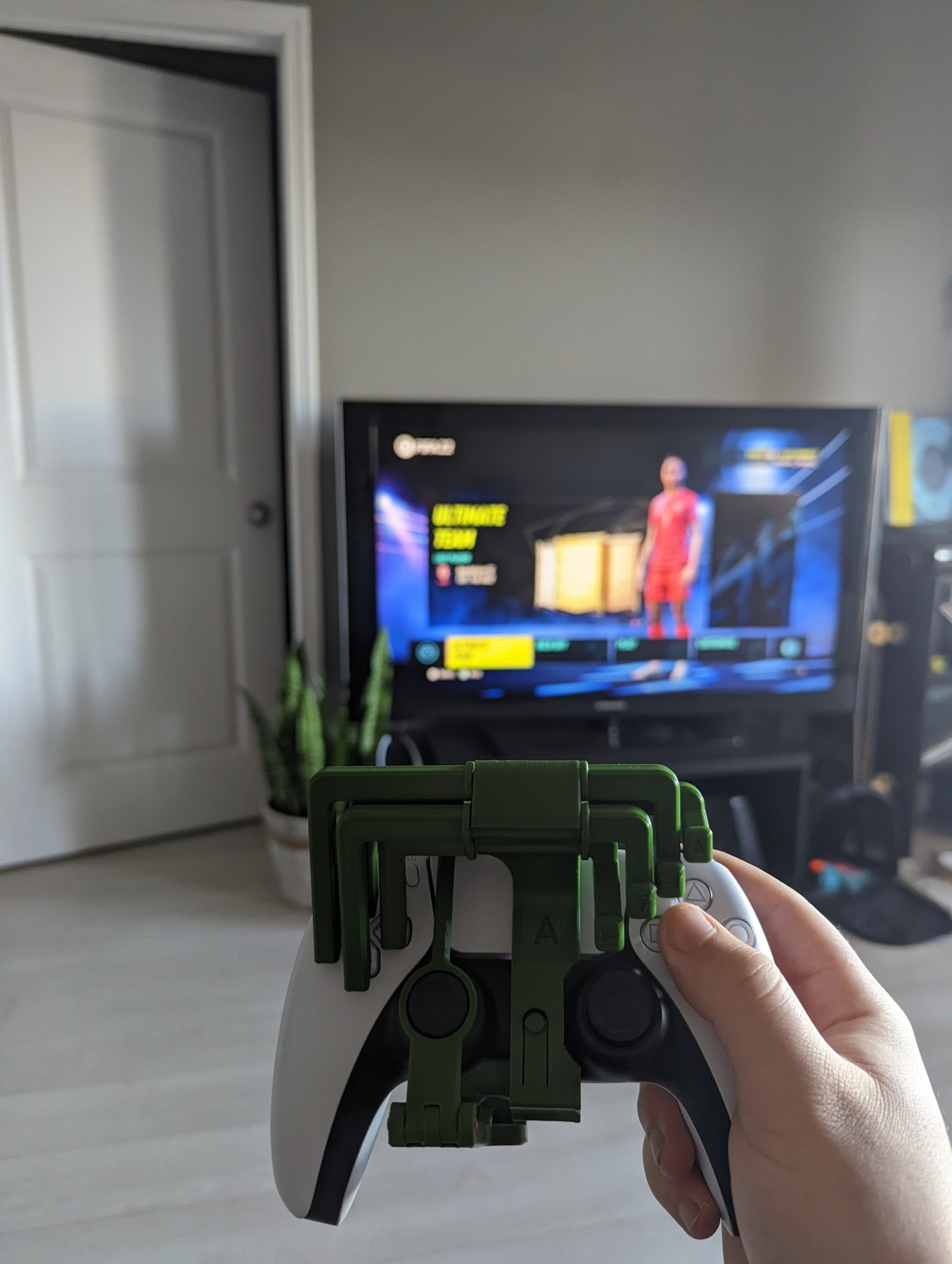 Holding a PS5 controller with a 3d printed controller mod attached. A FIFA video game is on the TV in the living room background.