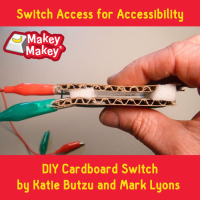 A hand holding a cardboard assistive switch with alligator clips attached.