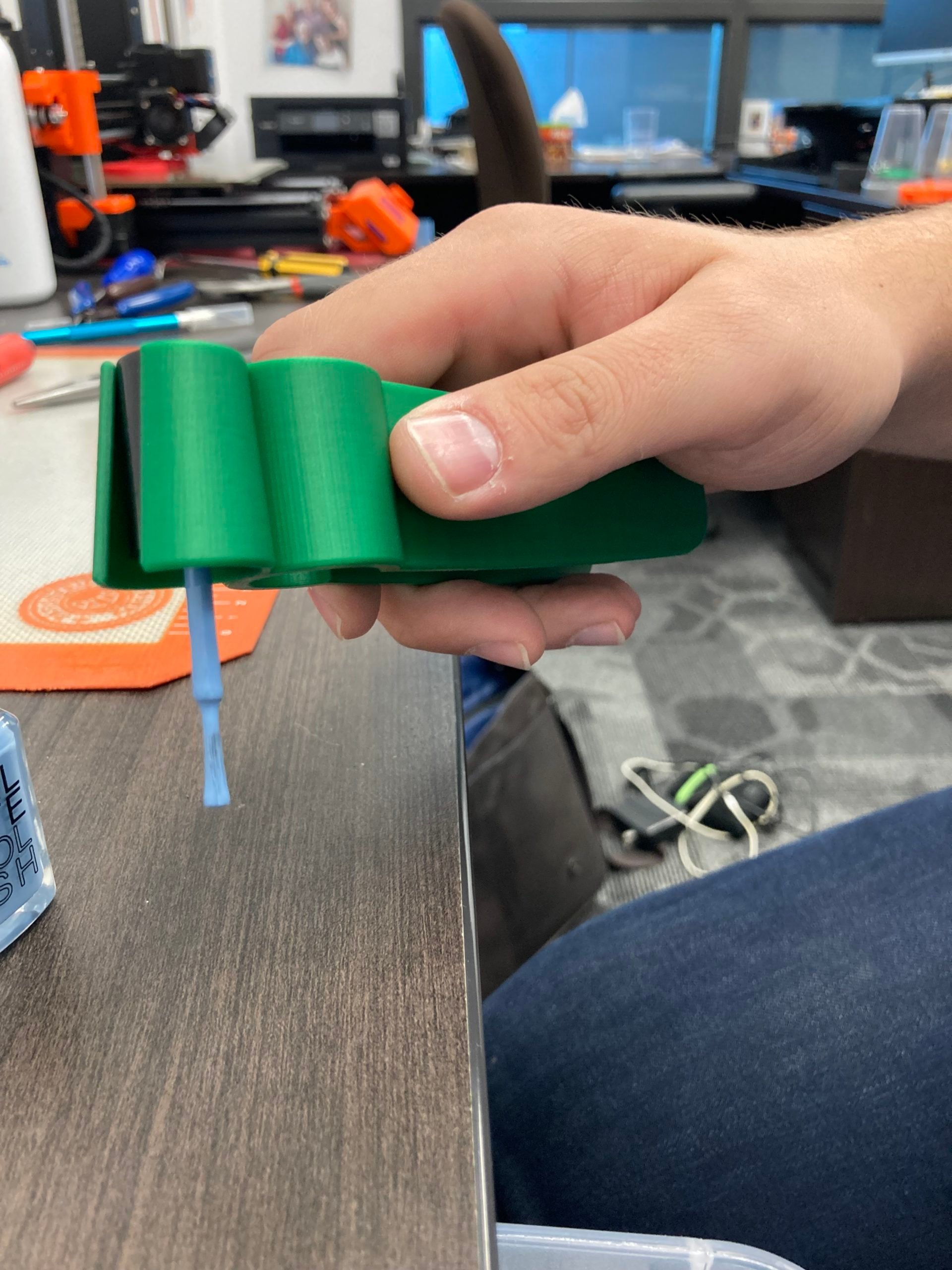 A hand holding a green 3d printed assistive nail polish applicator with a blue polish brush attached.