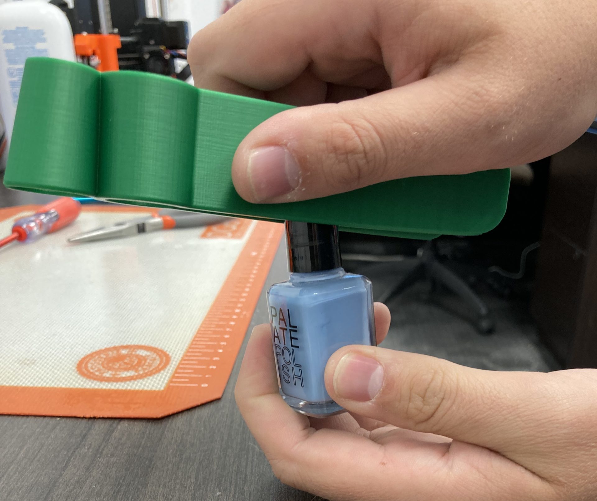 A hand using the handle of a green 3d printed assistive nail polish applicator to twist off the cap on a blue nail polish.