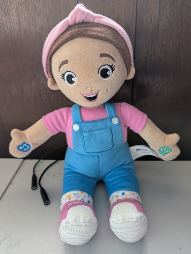 A plush Ms. Rachel doll wearing a pink shirt, blue overalls, and a pink headband sits upright on a table against a dark wood background. Two black 3.5 mm switch input jacks extend from the doll’s side, indicating it has been adapted for activation with external assistive switches.