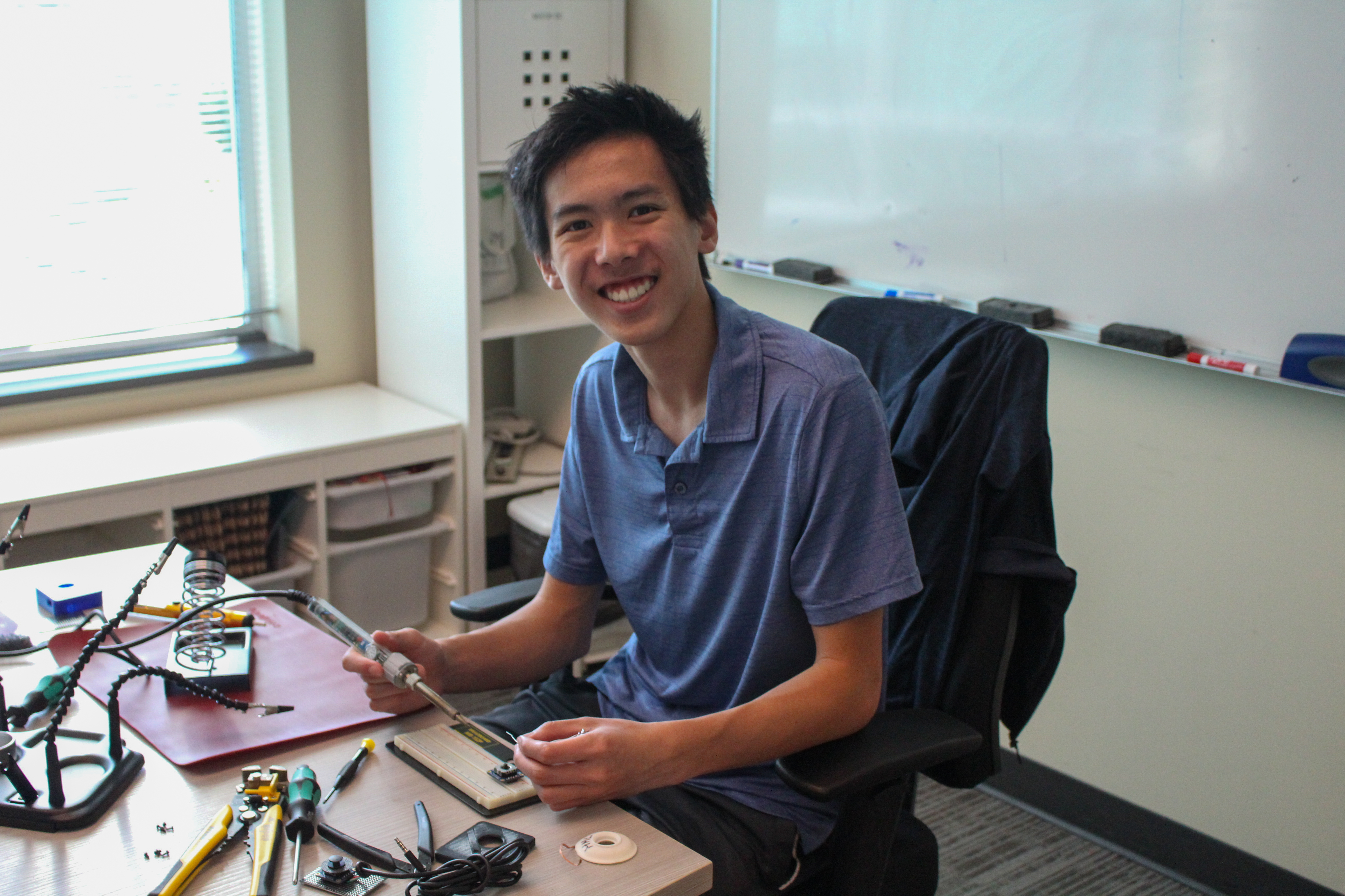 Ethan holds a soldering iron while he works on a device in an office boardroom.