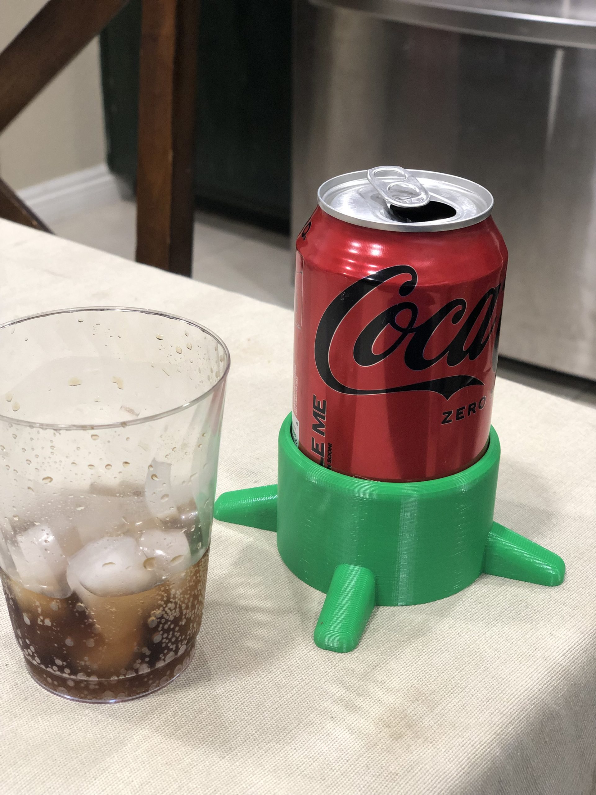 A can of coke sitting in a green 3d printed can stabilizer on a kitchen counter. To the left is a glass of coke and ice that's half full.