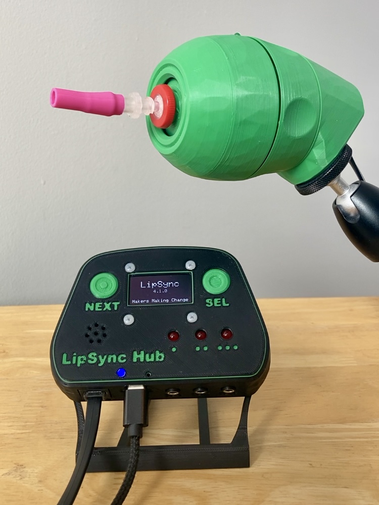 A green 3D printed LipSync joystick with a pink mouthpiece, mounted and shown above a black LipSync Hub on a table. On the Hub screen reads "LipSync v4.1.0 Makers Making Change."