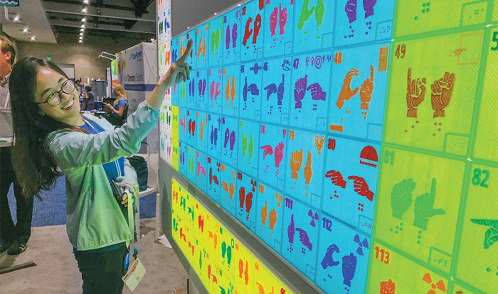 Picture of a woman interacting with a large 3d printed periodic table that has sign language and Braille