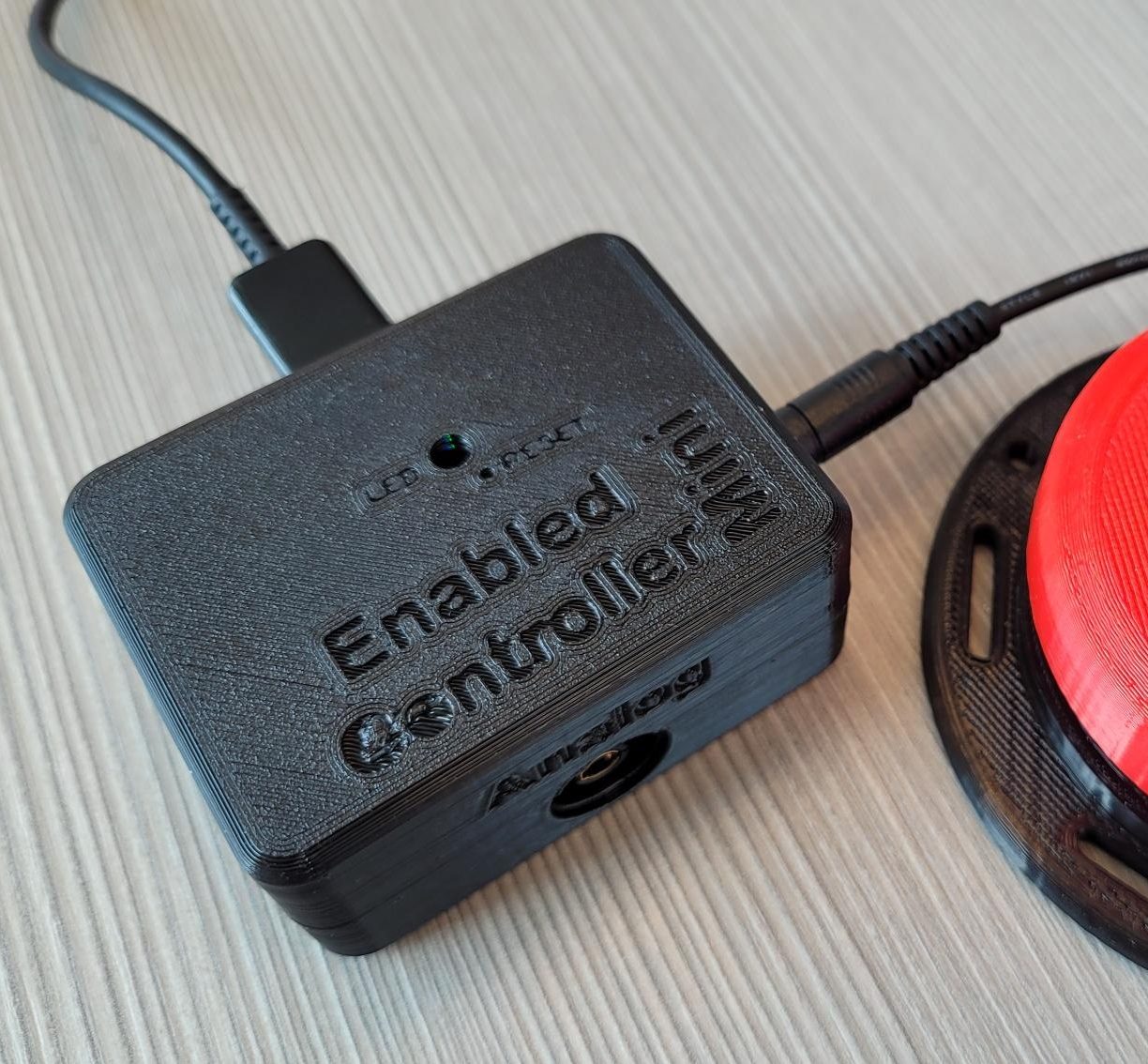 A picture of Enabled Controller Mini in a black 3D printed enclosure next to an adaptive switch in red color.