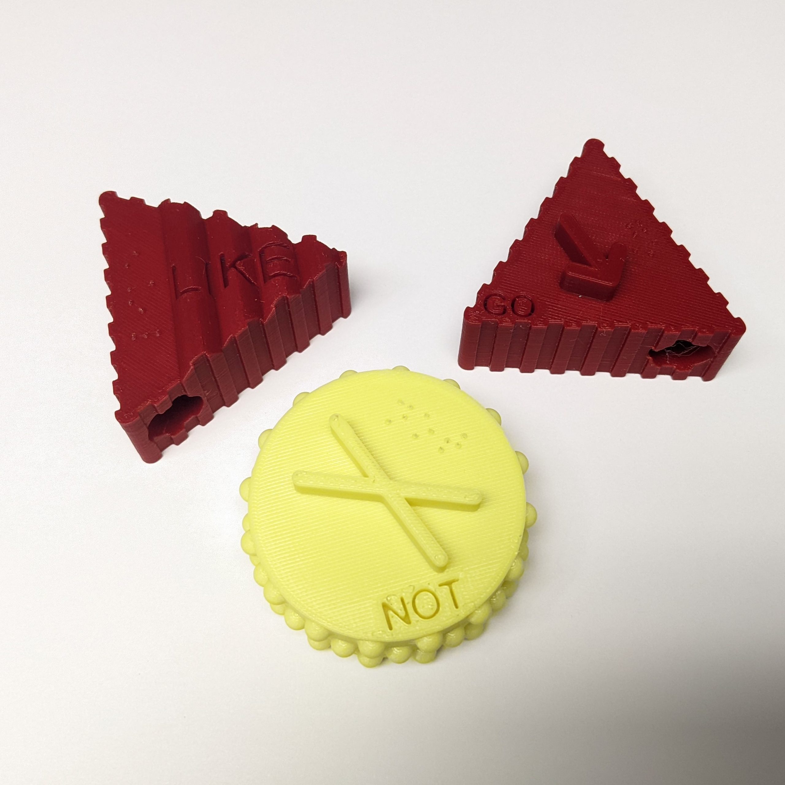 Two red and one yellow 3d printed tactile symbols against a white background. There are three symbols, a triangular red 'Go', a triangular red 'Like', and a circular yellow 'Not'.
