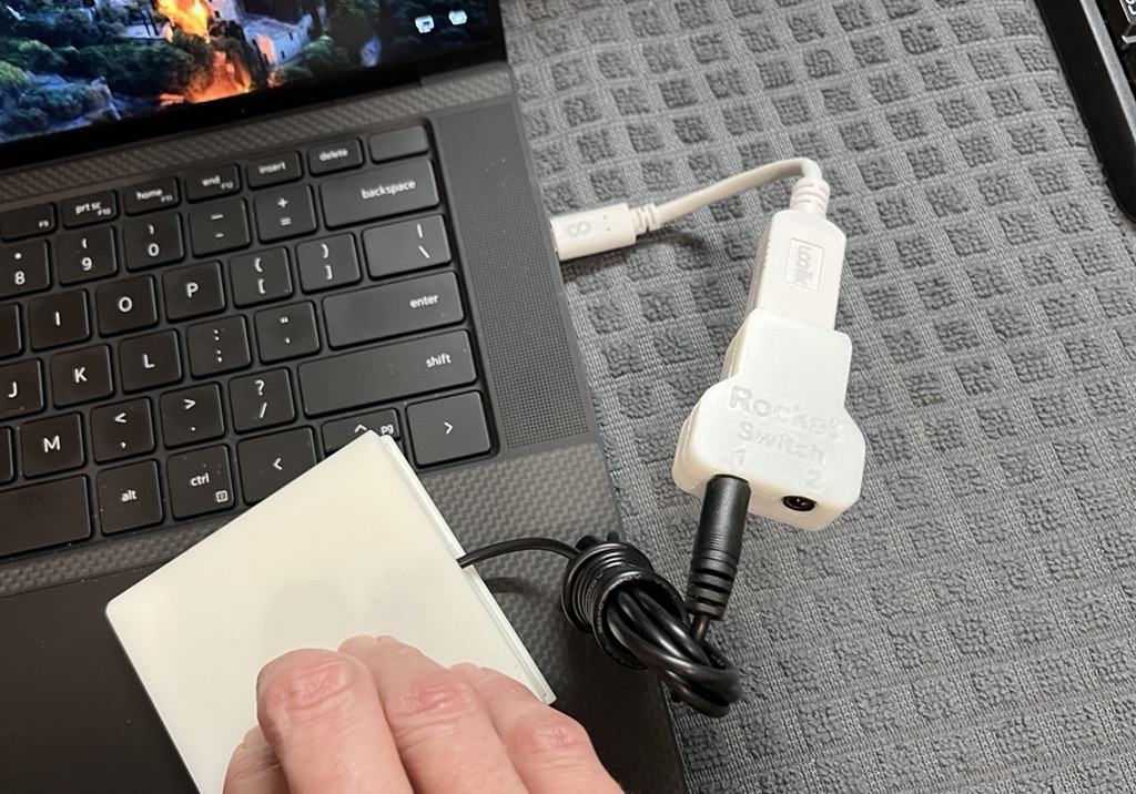 A hand pressing on a white assistive switch plugged into the Rocket Switch, which is plugged into a black laptop.