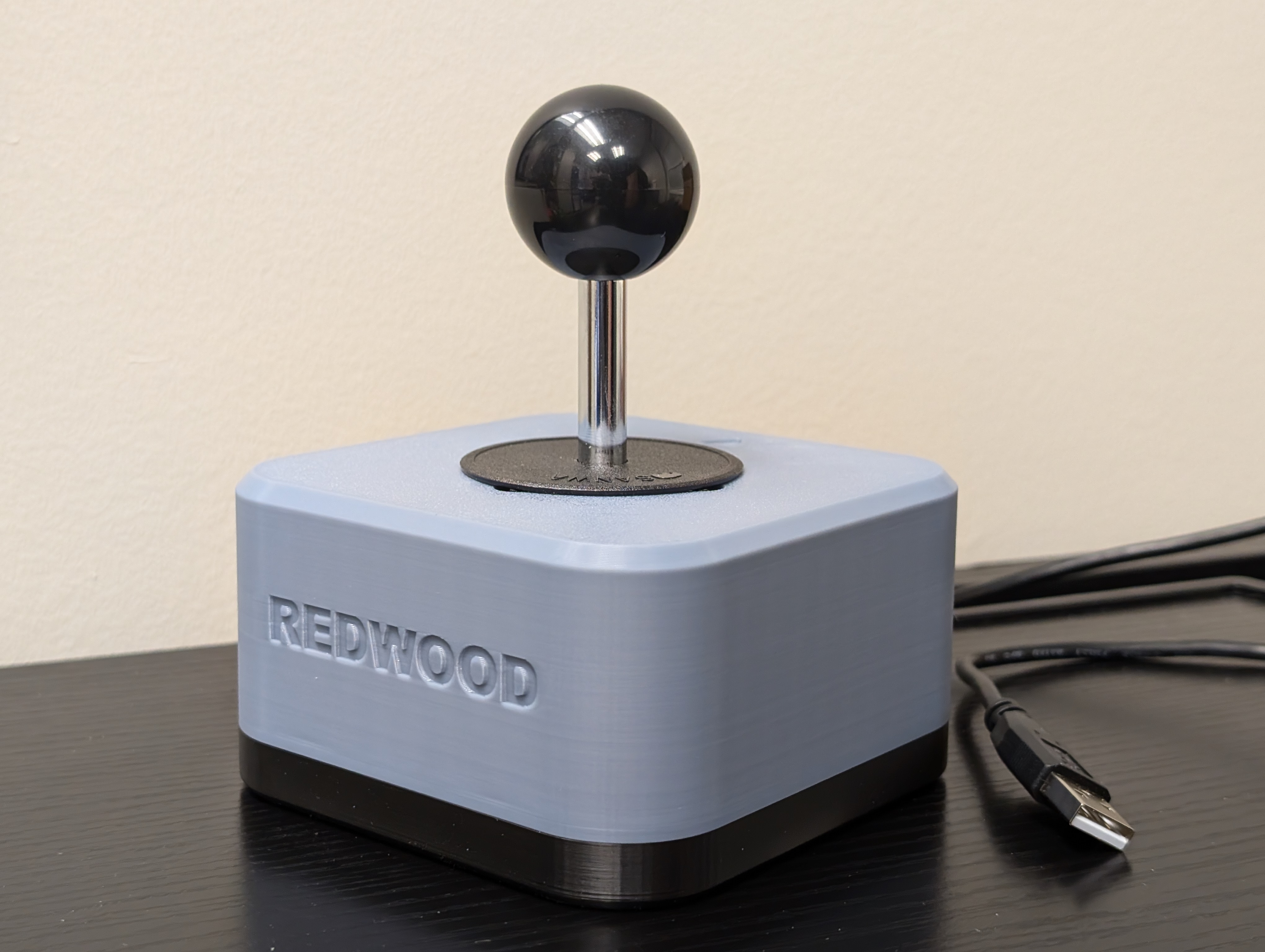 Isometric view of the Redwood joystick with a glossy black ball topper. A USB type A cable can be seen on the side. Top enclosure is light blue and base is black.