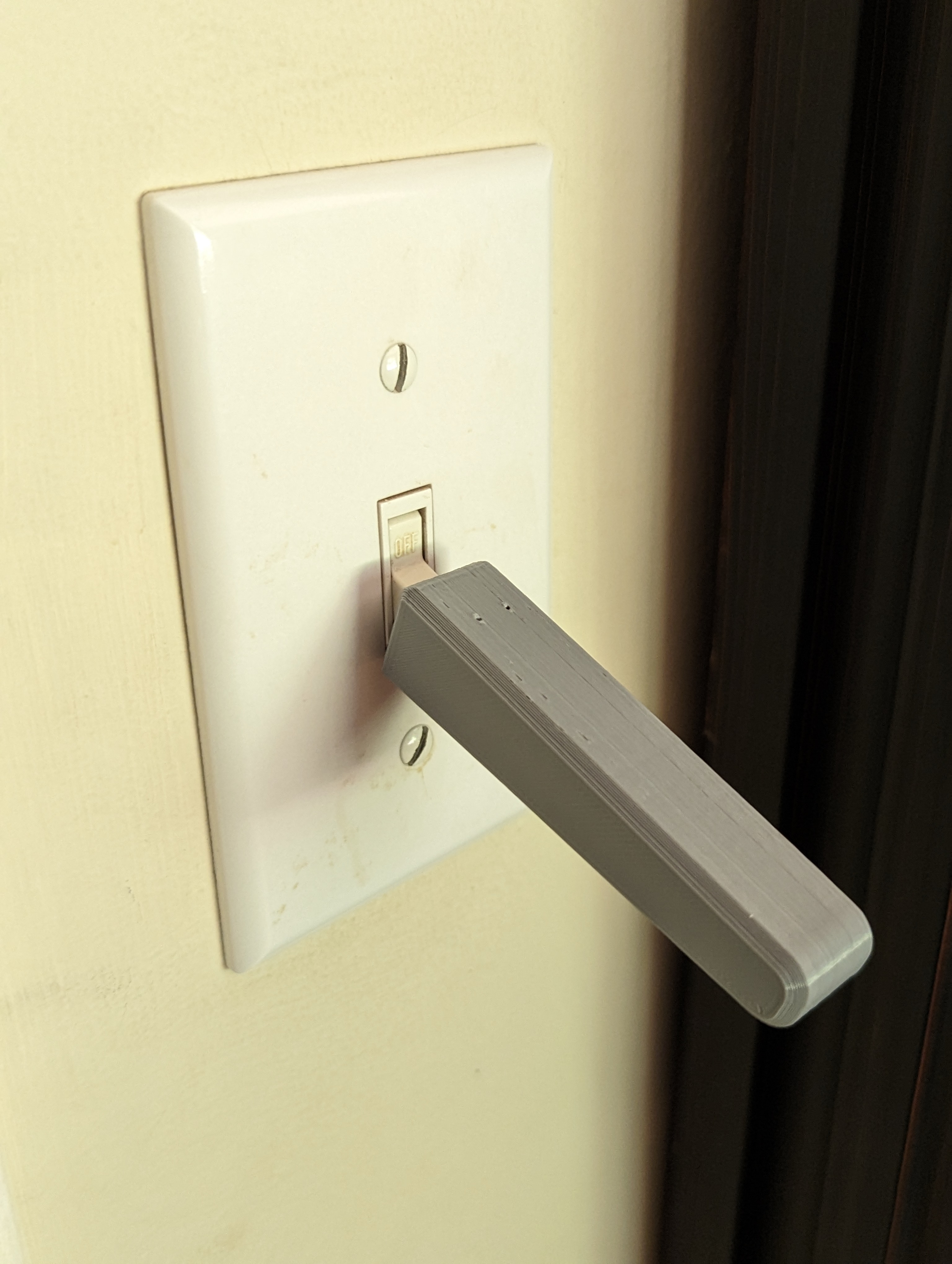 A grey light switch extension lever arm install over a standard light switch against a light tan coloured wall.
