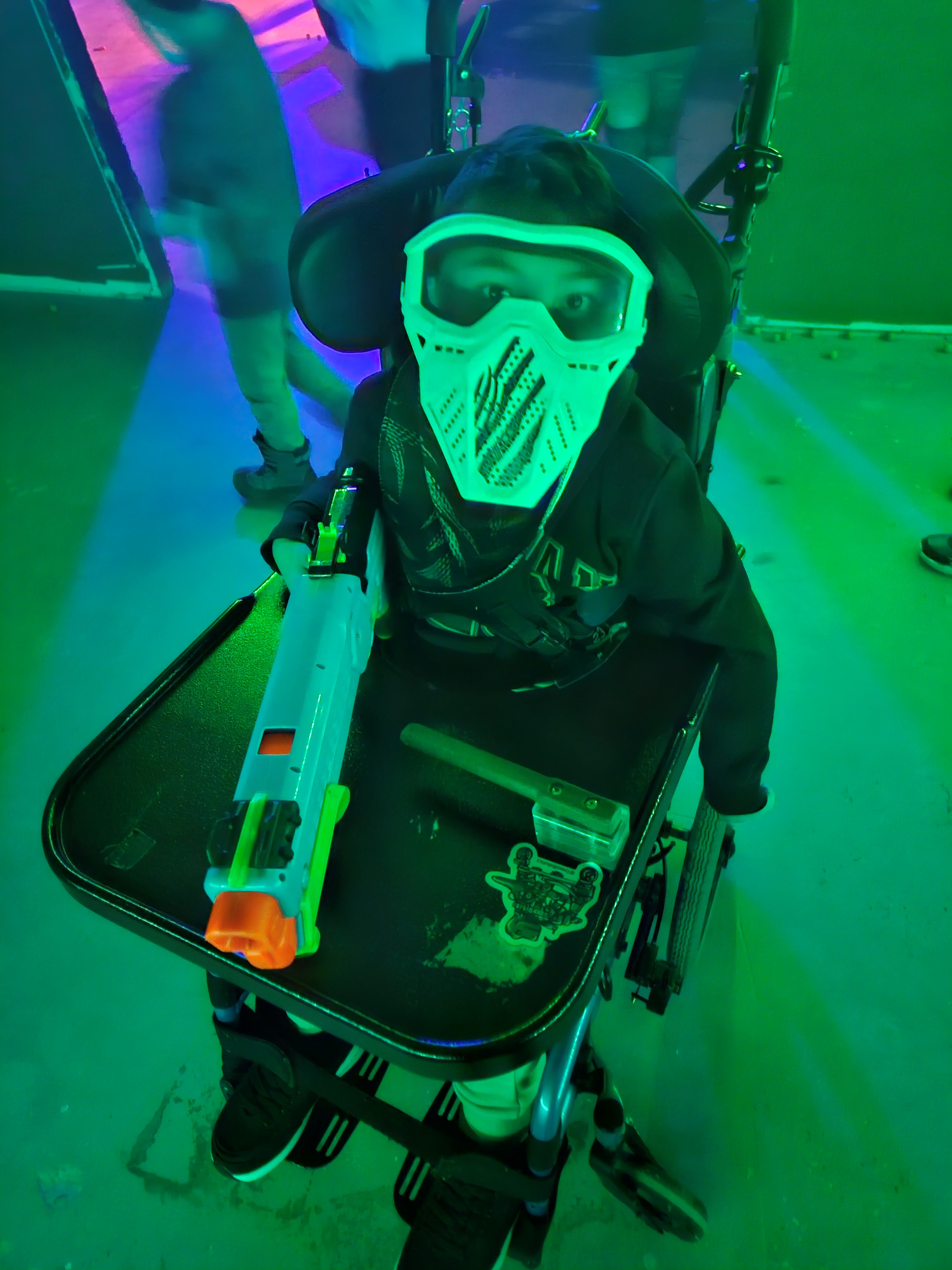 Adam in his wheelchair in a neon-lit nerf facility with a switch-adapted nerf gun.