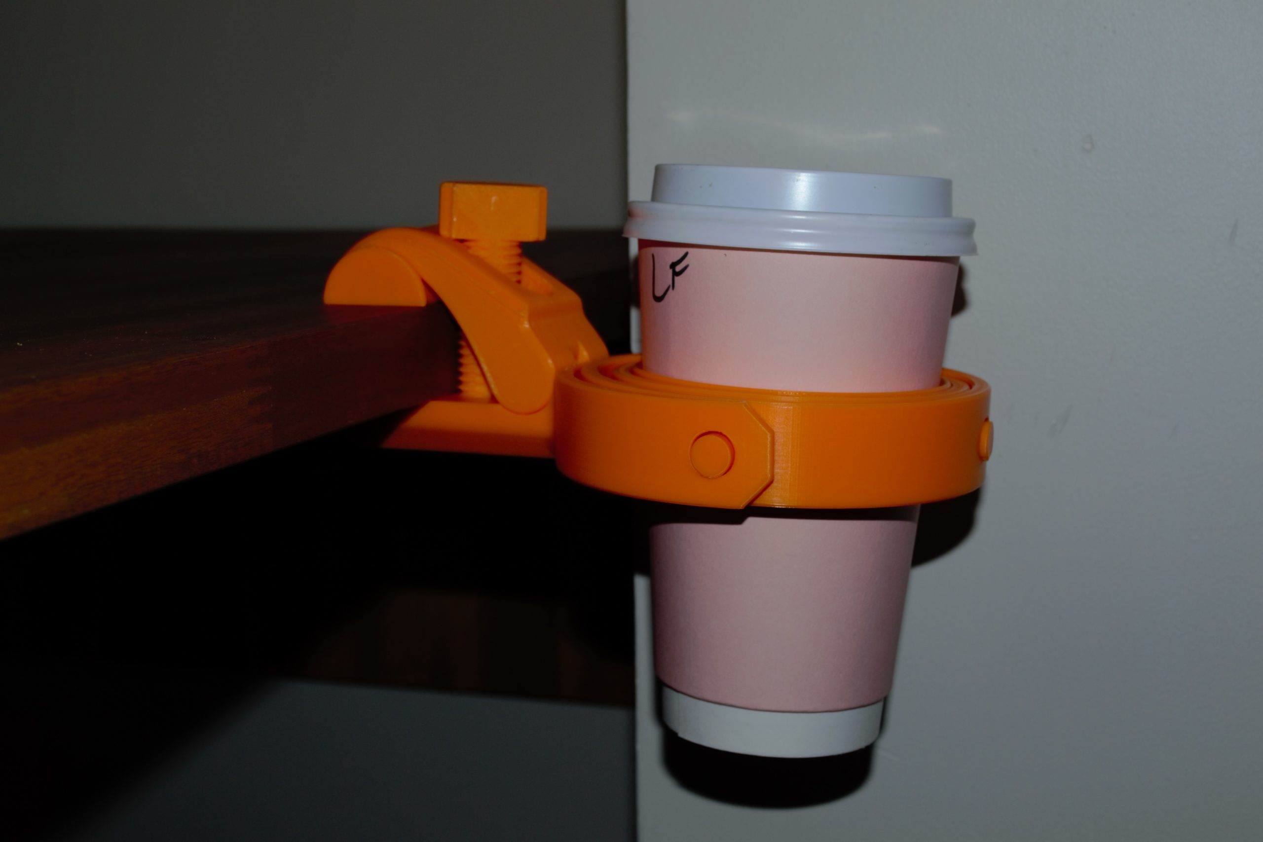 An orange 3d printed travel coffee gimbal clamped to a flat edge and holding a disposable coffee cup..