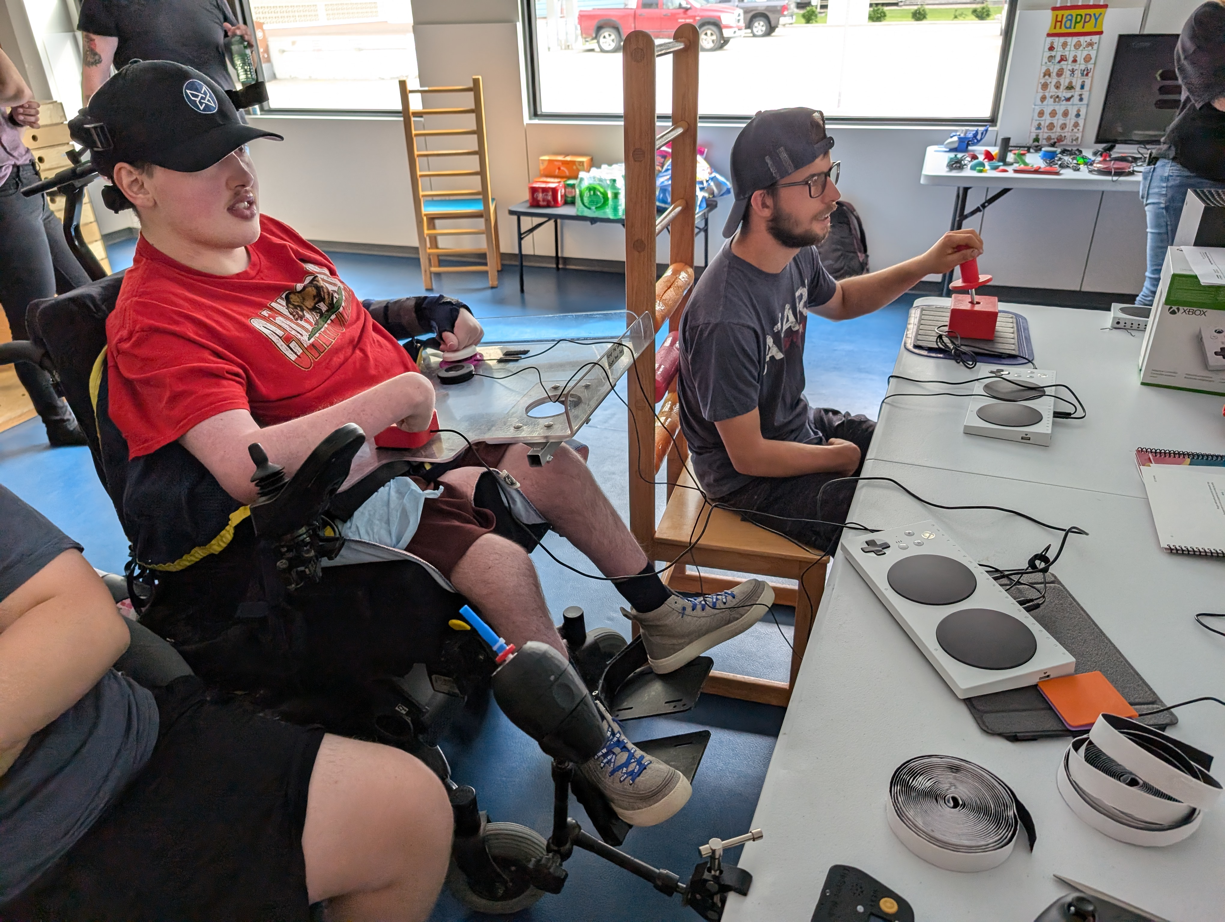 Two gamers, including one in a wheelchair, use adaptive joysticks and switches to play a game.