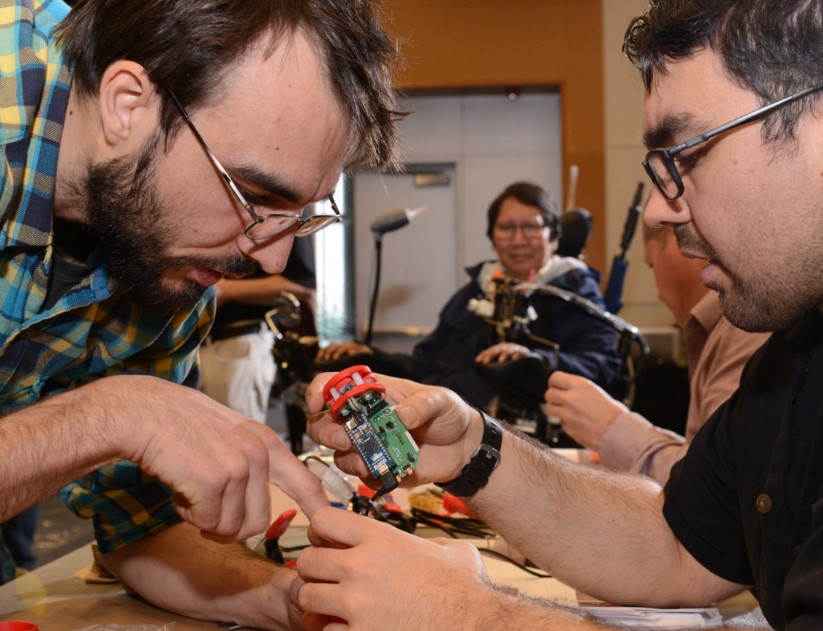 Jake and another maker work on a LipSync at the BC Tech Summit as a person in a wheelchair looks on.