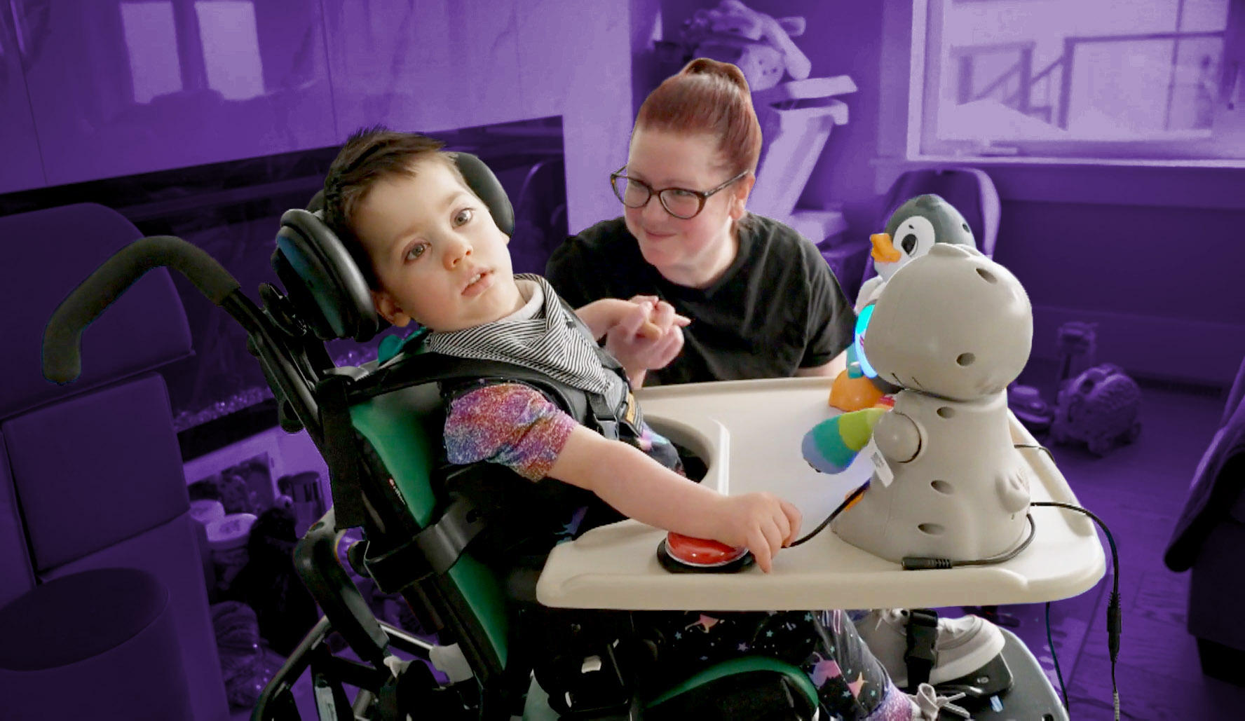 A child with a disability in a wheelchair plays with a switch-adapted toy while a clinician looks on.