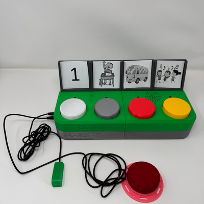 The Chatterbox with four labels printed paper labels to represent the messages that could be on each button. The Chatterbox is a green rectangle with four circular buttons, and the label holders are squares on the back at an angle. The printed labels are from left to right: the number one; a child raising their hand; a school bus; and three children singing.