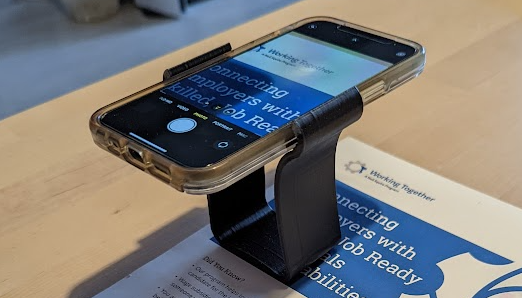 A black 3d printed magnification stand holding an iphone. The camera app is opened and zoomed in on a paper underneath.