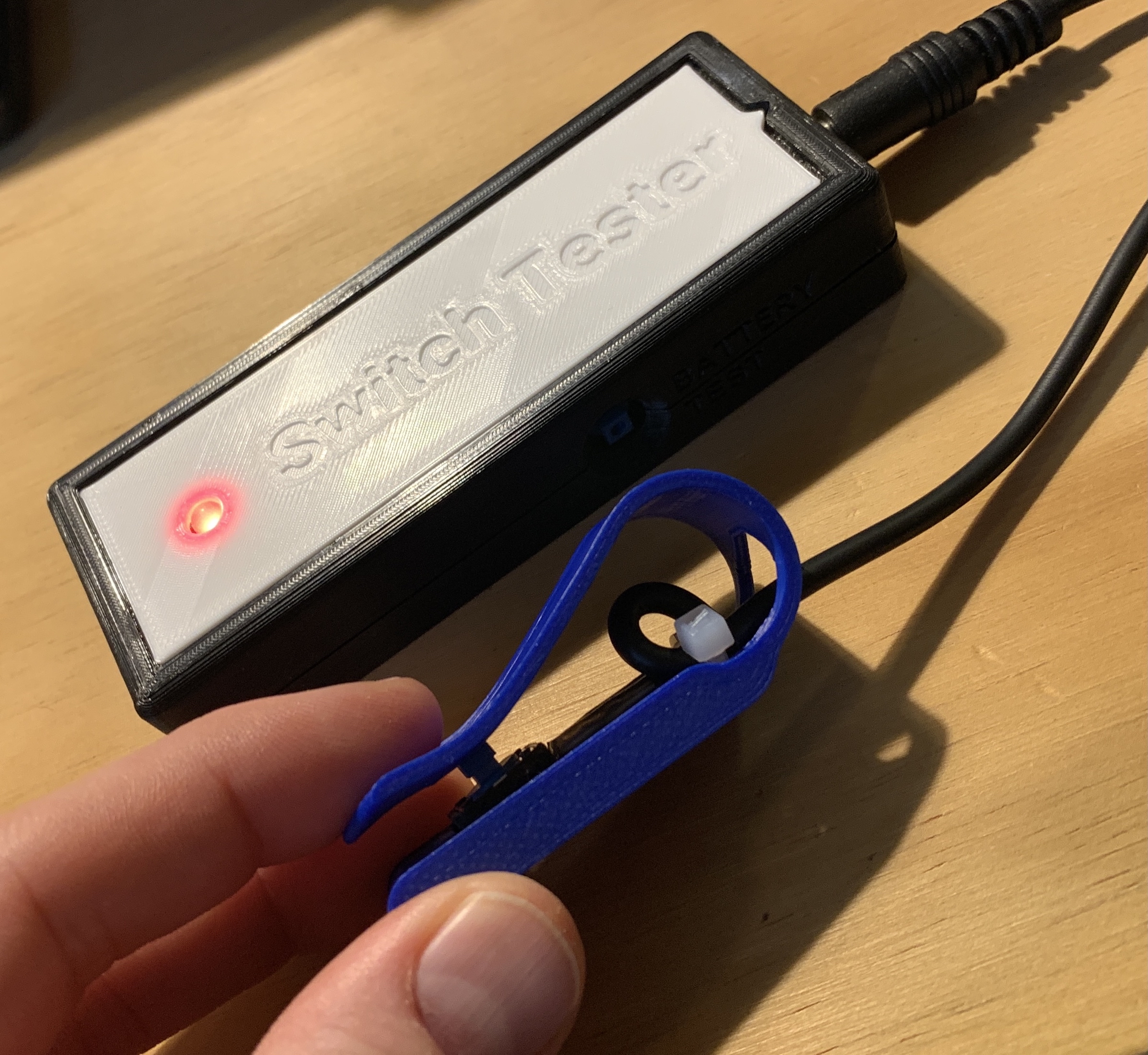 A hand pressing down on a blue 3d prnited solderless switch plugged into a blakc and white switch tester. The red lLED on the tester is lit up.