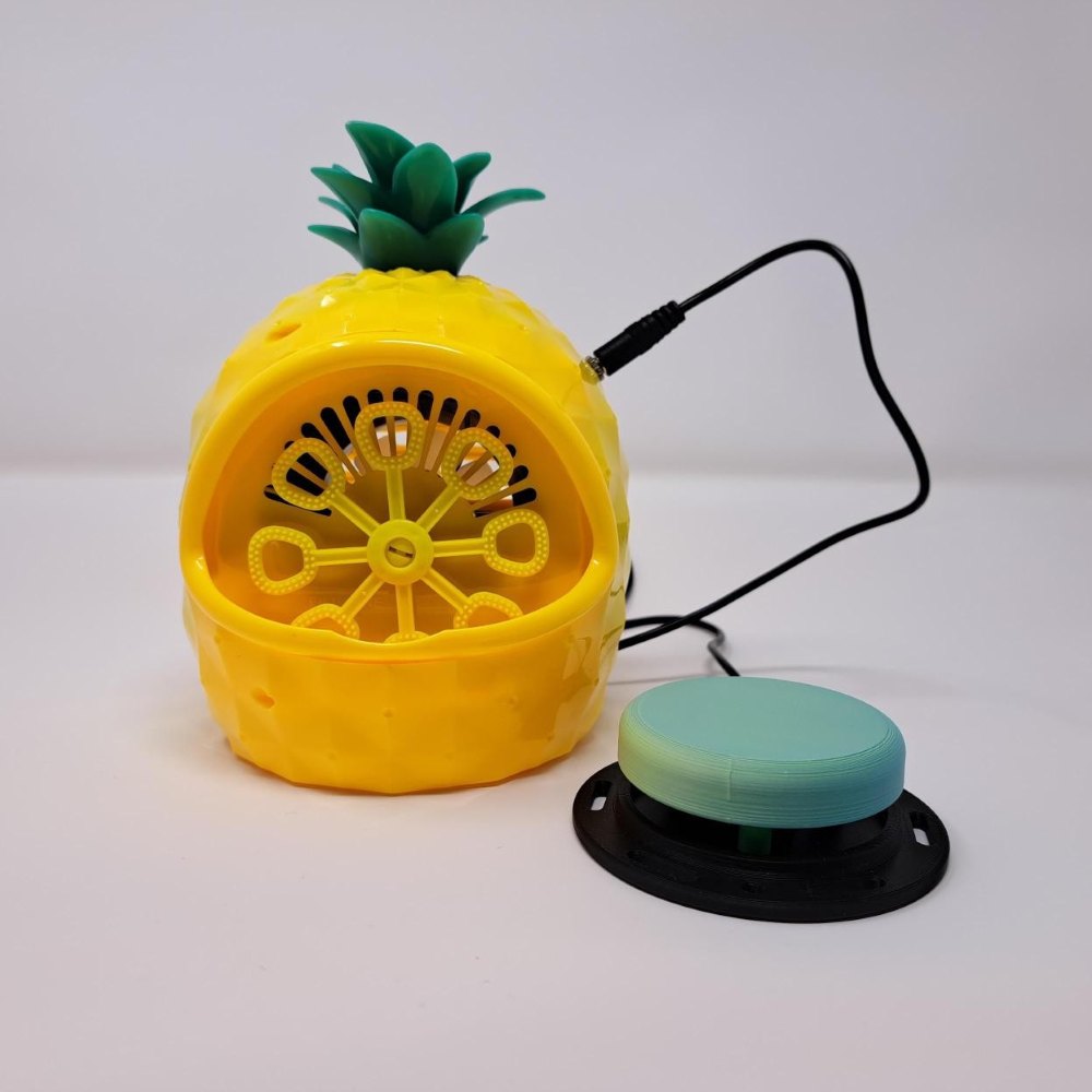 A yellow pineapple bubble machine that has been switch adapted to work with an assistive switch. A large green assistive switch is plugged into it.