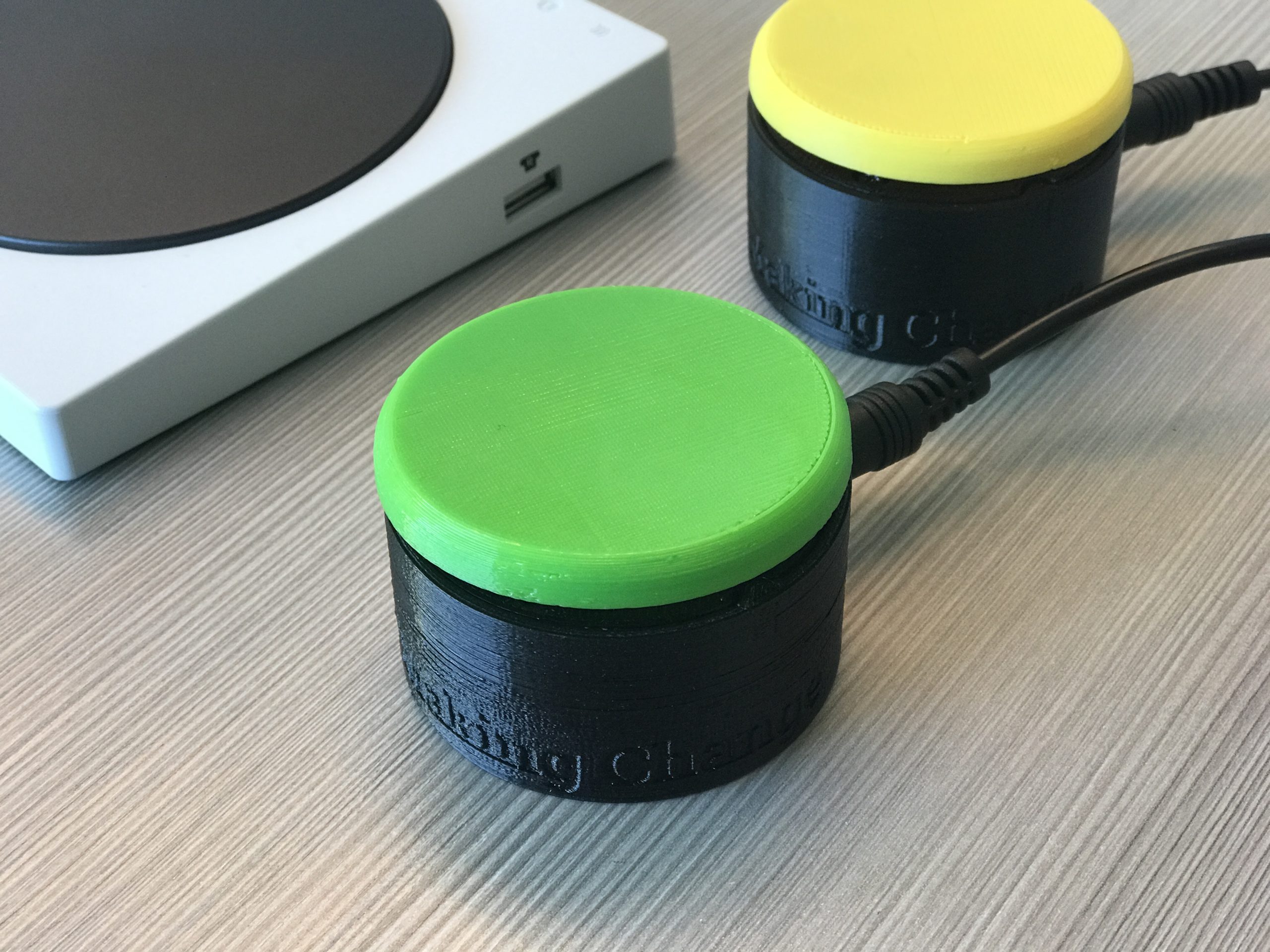 Picture of two MMC60 switches. One has a green top and one has a yellow top. They are pictured next to a Microsoft Xbox Adaptive Controller.