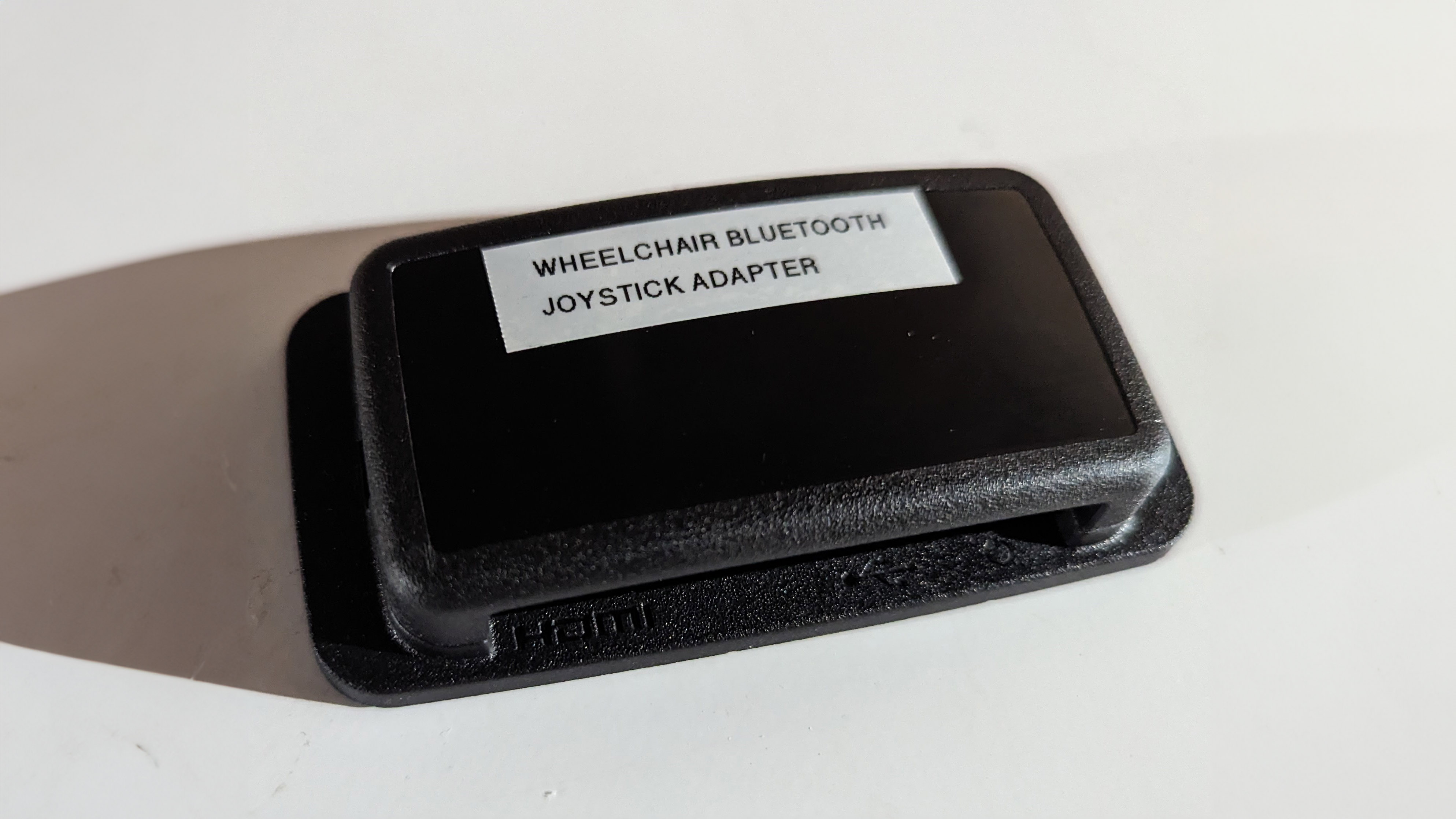 A black buckle with a white tape on top that says "Wheelchair Bluetooth Joystick Adapter"
