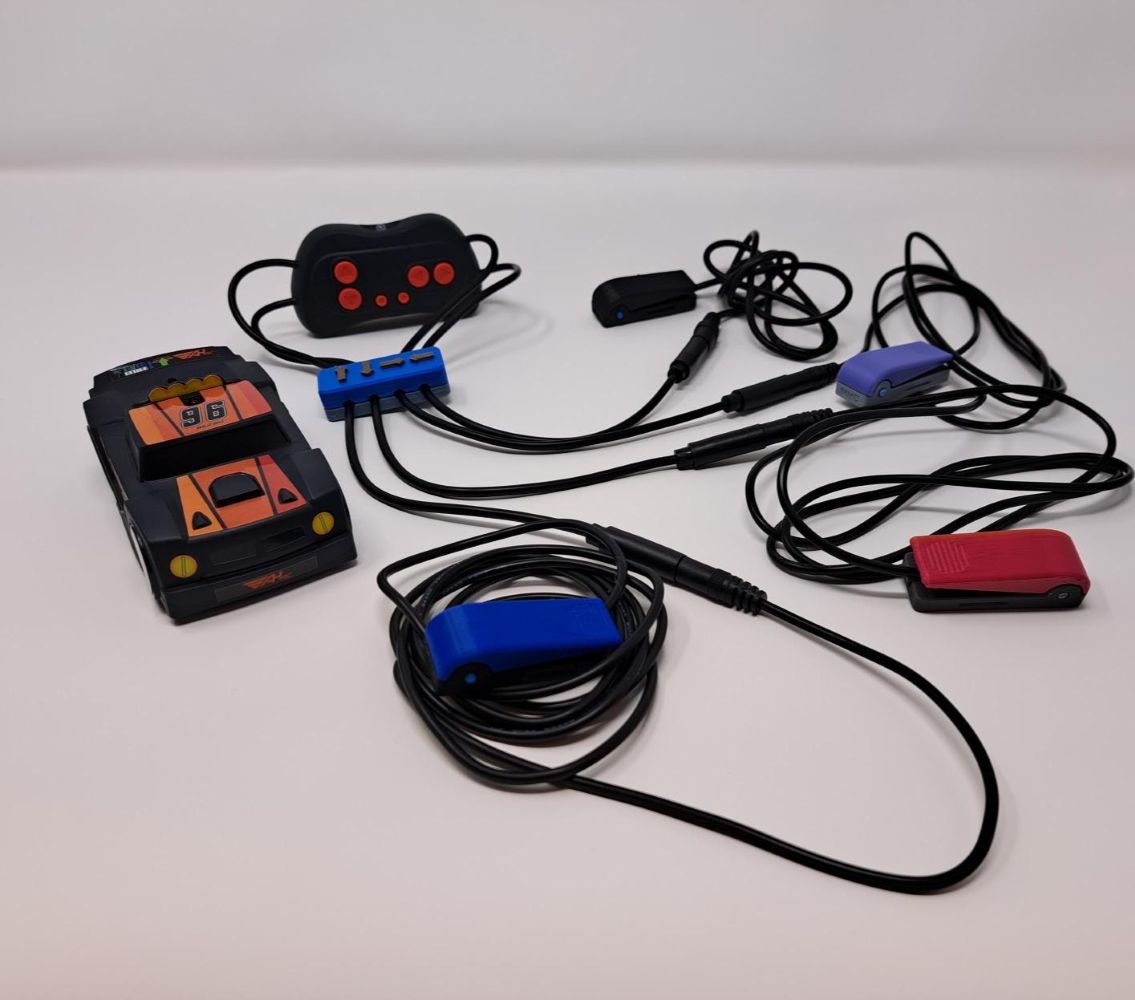 A black and orange remote controlled car sits next to its remote controller, which has been adapted to be used with assistive switches. Four assistive switches are plugged into the remote.