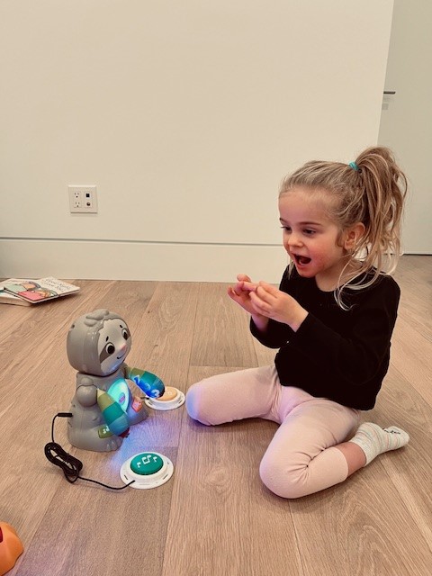 A young girl in a black shirt and pink leggings playing with a switch adapted toy sloth.